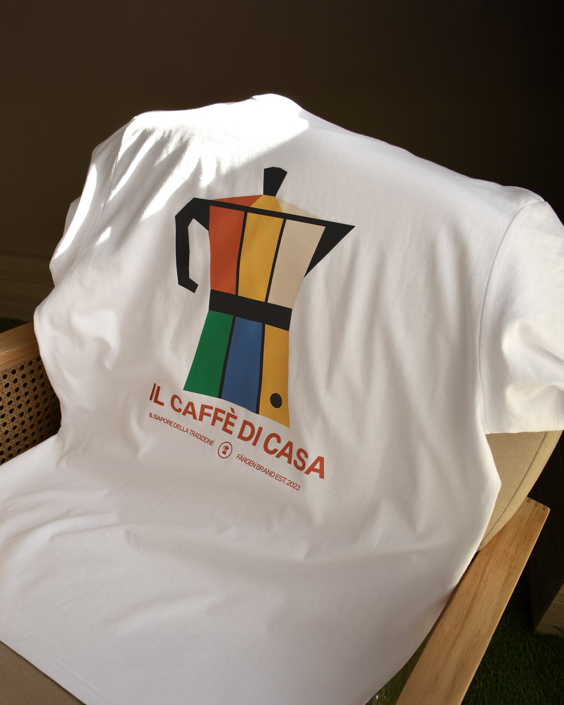 Camiseta blanca Färgen Il Caffè di Casa doblada sobre una silla mostrando el diseño trasero con cafetera italiana y texto Il Caffè di Casa.