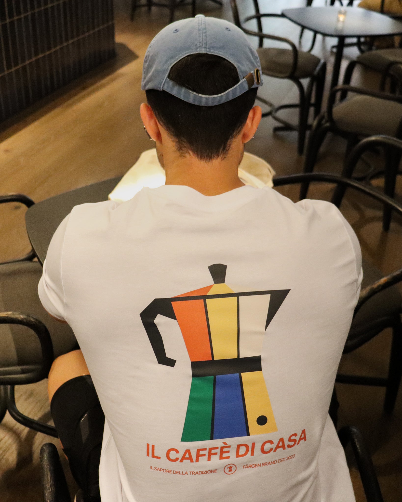 Hombre con camiseta blanca Färgen Il Caffè di Casa sentado en cafetería mostrando el diseño trasero con cafetera italiana de colores estilo Bauhaus.