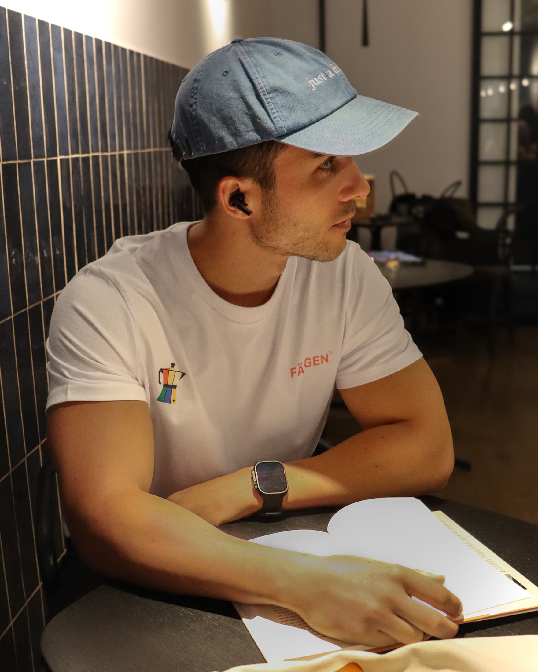 Hombre con camiseta blanca Färgen Il Caffè di Casa mostrando el diseño delantero con cafetera de colores y logotipo Färgen. Gorra just a chill guy
