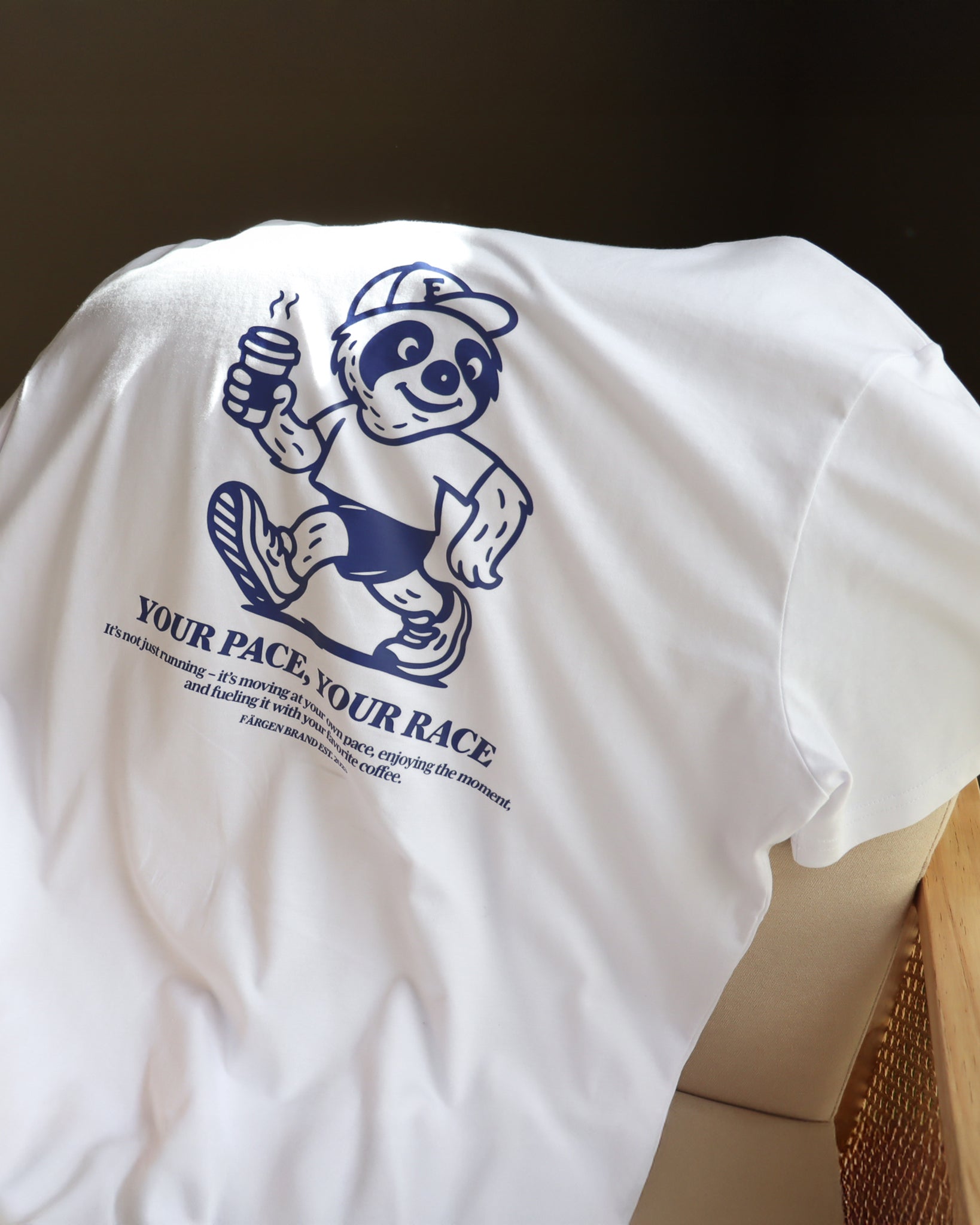 Camiseta Färgen Your Pace Your Race doblada sobre una silla mostrando el diseño trasero con perezoso corriendo y texto en azul.