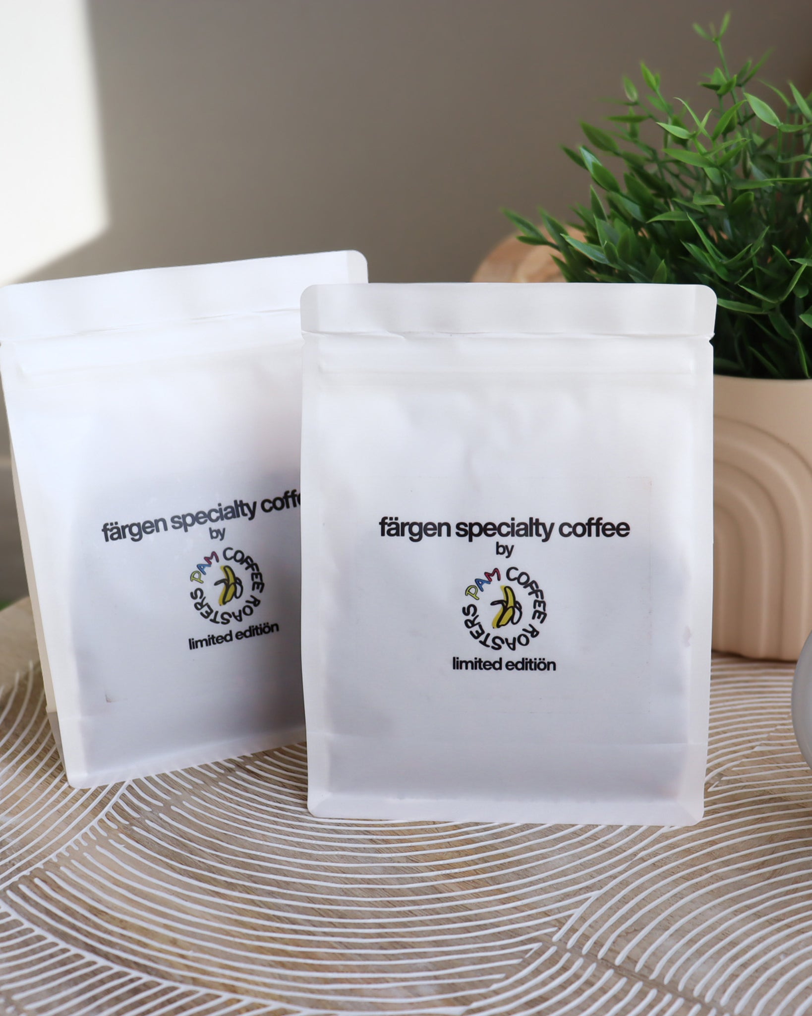 Dos bolsas de Färgen Specialty Coffee x Pam Coffee Roasters edición limitada junto a una planta y una taza, sobre mesa decorativa.