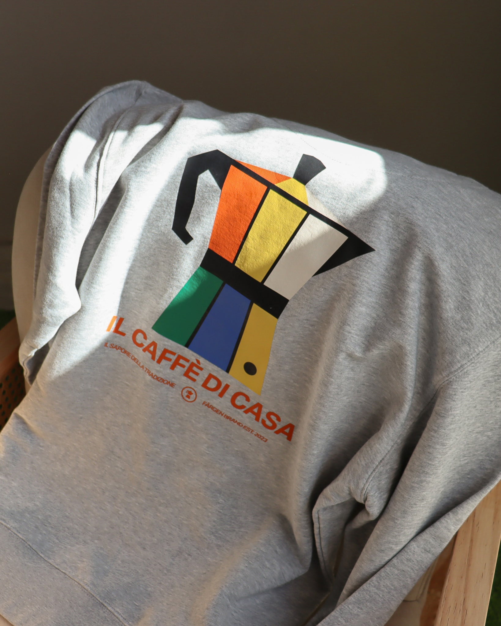 Sudadera gris Färgen Il Caffè di Casa doblada sobre silla mostrando el diseño trasero con cafetera italiana y texto Il Caffè di Casa.