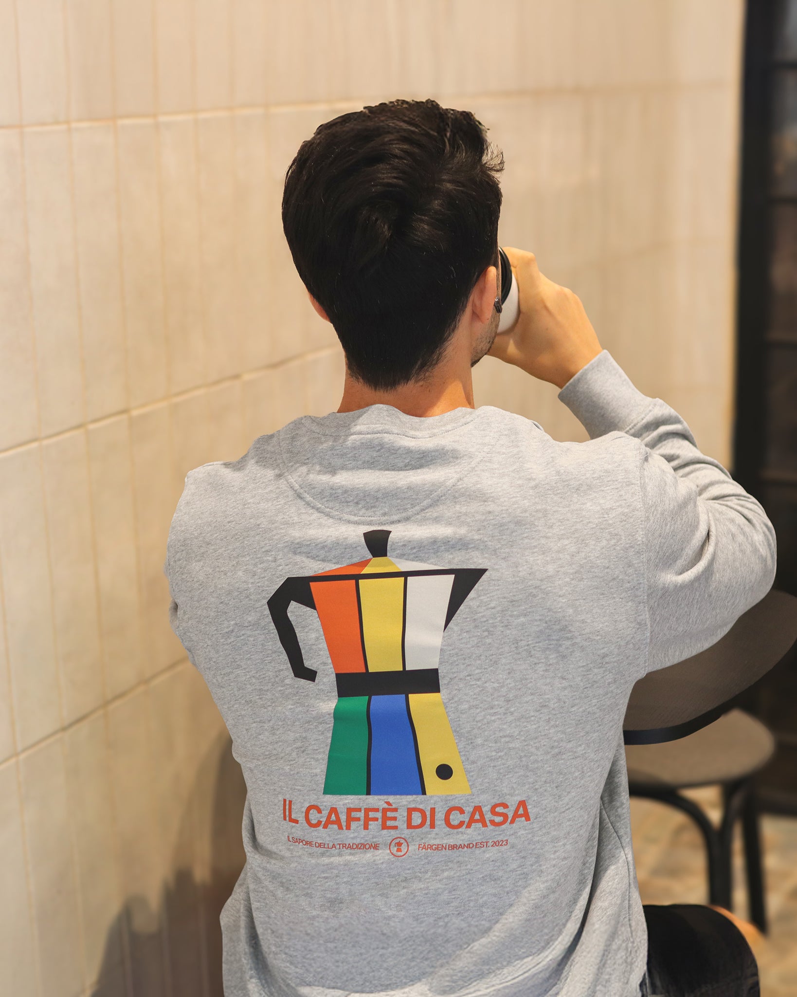 Modelo con sudadera gris Färgen Il Caffè di Casa de espaldas, mostrando el diseño trasero con cafetera italiana de colores Bauhaus y texto Il Caffè di Casa.