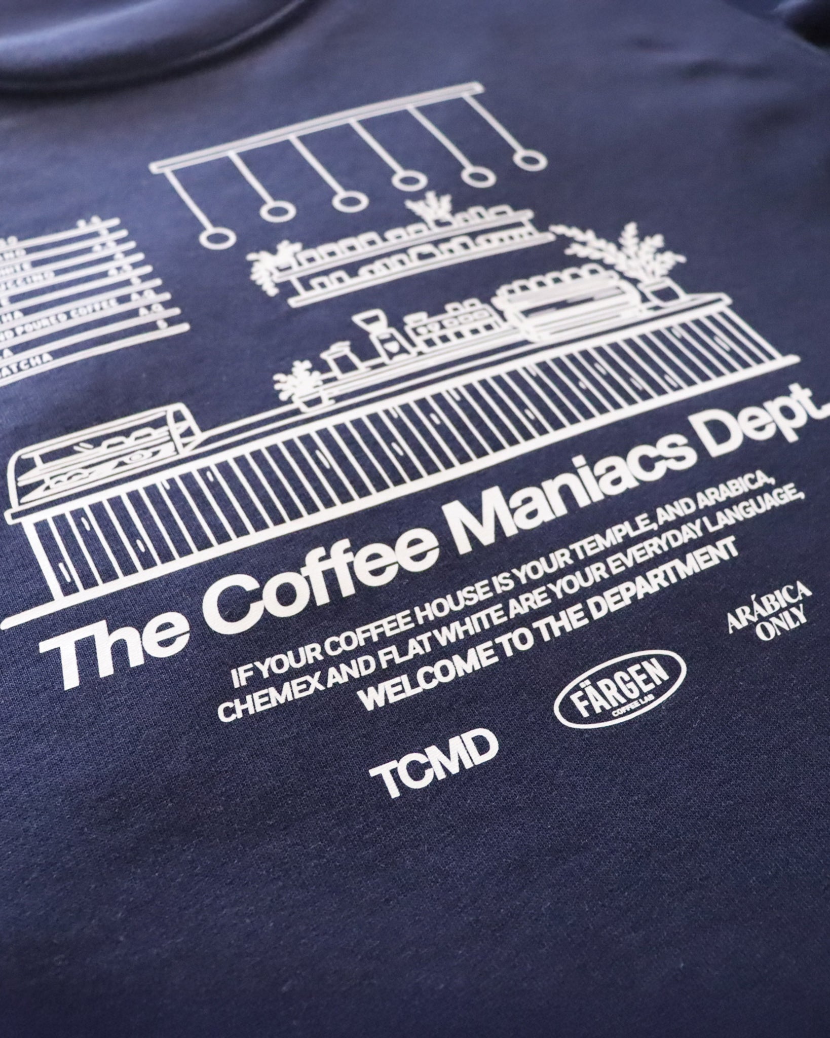 Detalle del diseño trasero de la sudadera Färgen The Coffee Maniacs Dept con ilustración de barra de cafetería y texto en blanco.