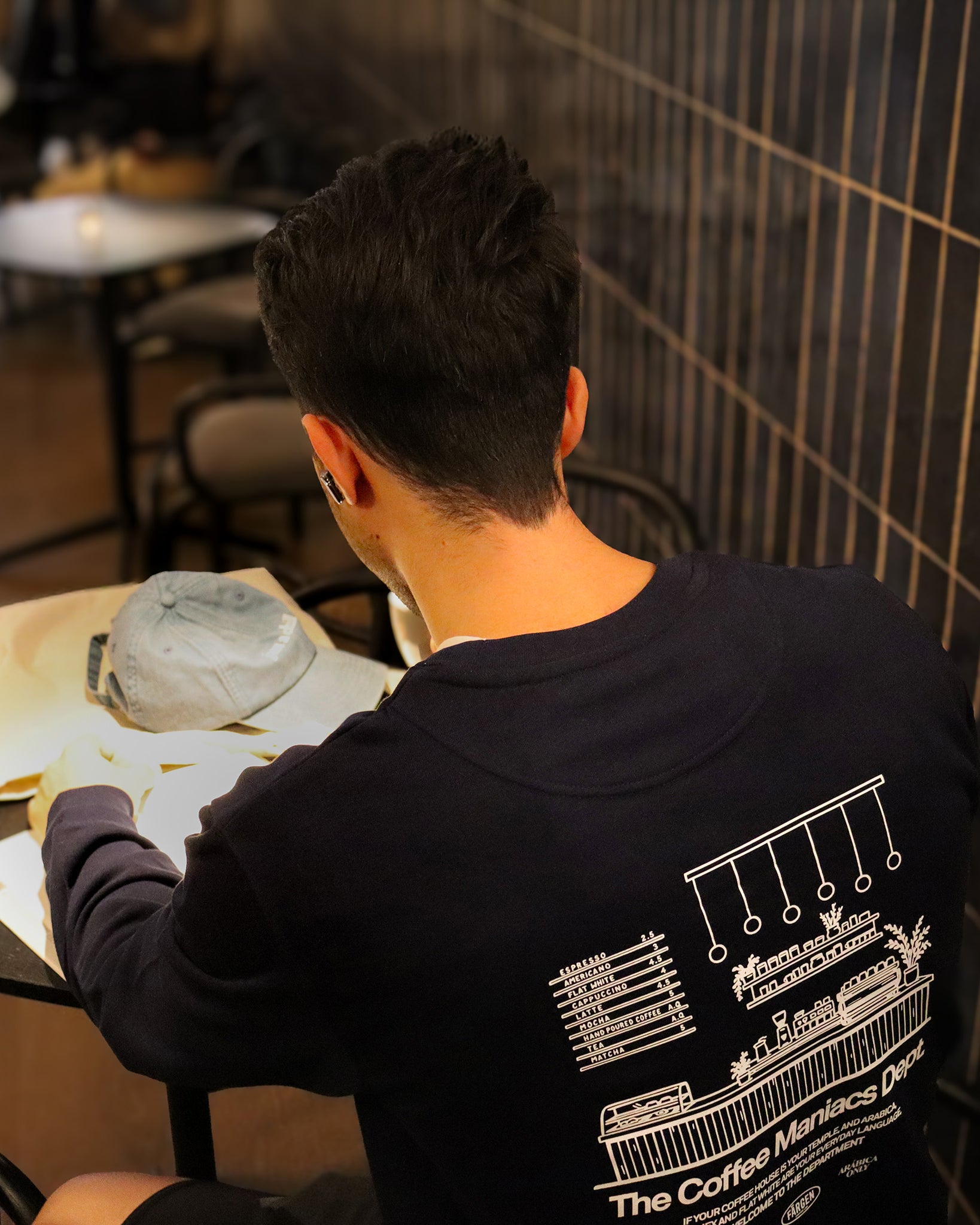Hombre con sudadera azul marino Färgen The Coffee Maniacs Dept sentado en una cafetería mostrando el diseño trasero con ilustración de barra de café.
