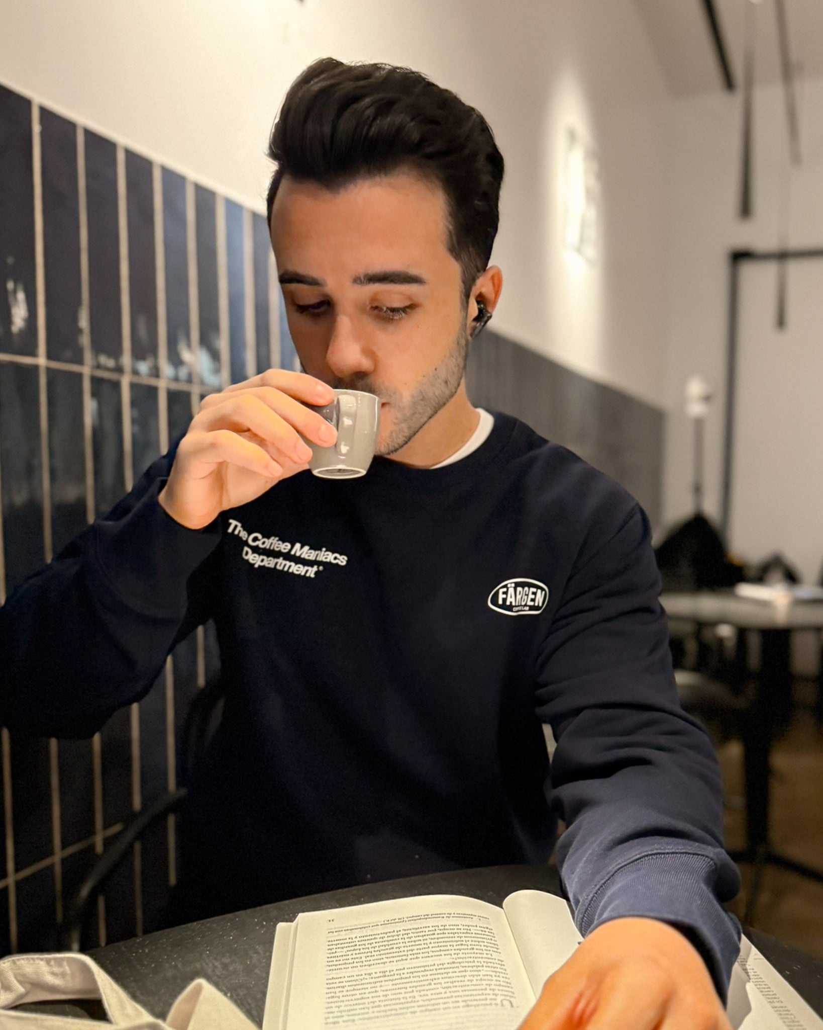 Hombre con sudadera azul marino Färgen The Coffee Maniacs Dept tomando café en una cafetería, mostrando el diseño delantero con logotipo Färgen Coffee Lab.