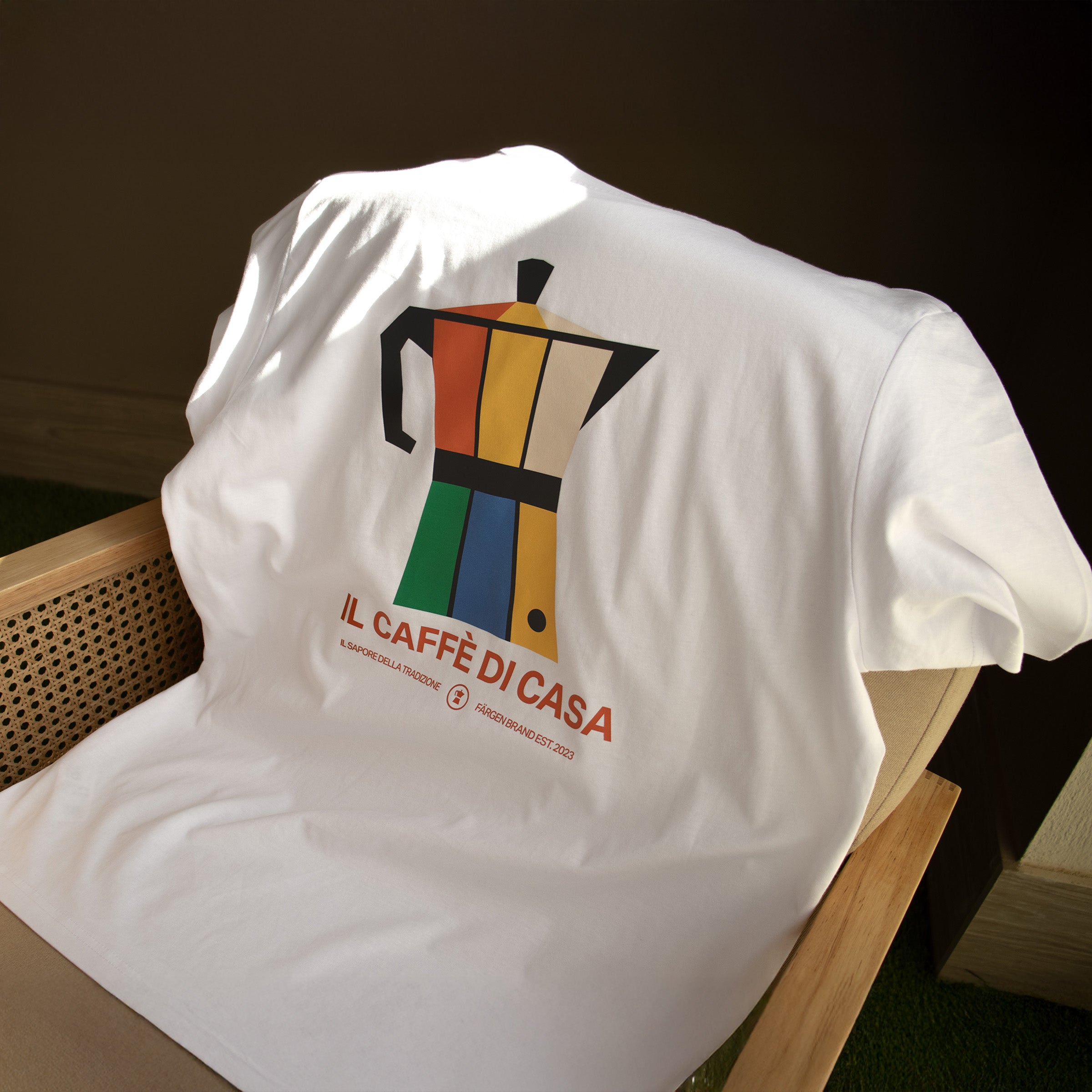 Camiseta blanca Färgen Il Caffè di Casa doblada sobre una silla mostrando el diseño trasero con cafetera italiana y texto Il Caffè di Casa.