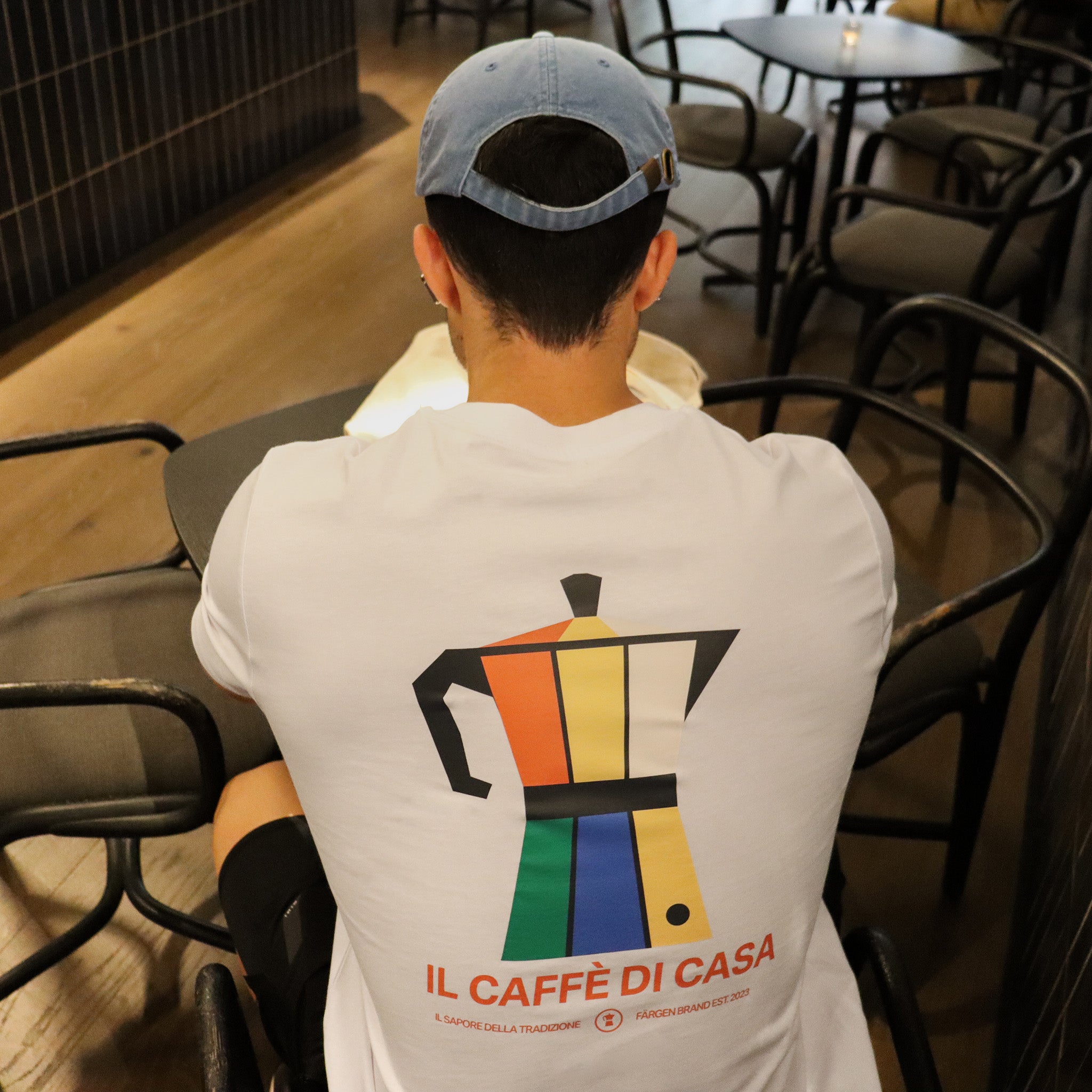 Hombre con camiseta blanca Färgen Il Caffè di Casa sentado en cafetería mostrando el diseño trasero con cafetera italiana de colores estilo Bauhaus.