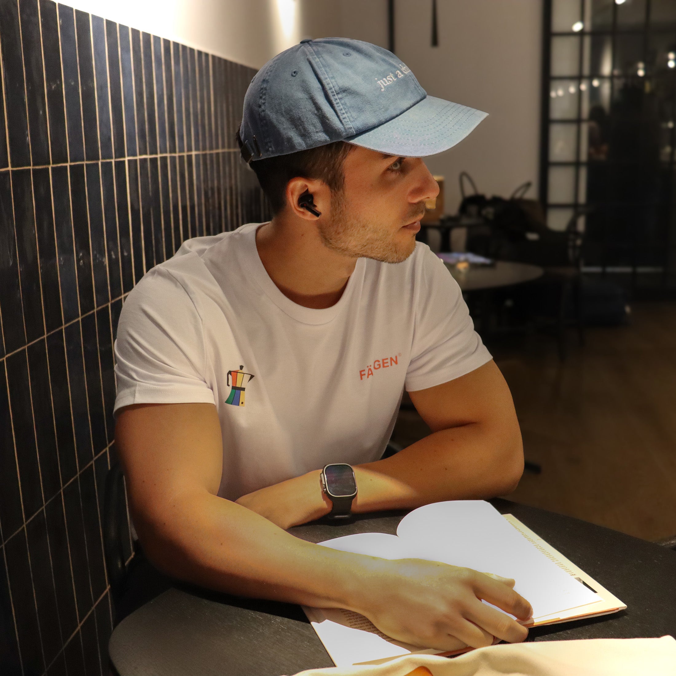 Hombre con camiseta blanca Färgen Il Caffè di Casa mostrando el diseño delantero con cafetera de colores y logotipo Färgen. Gorra just a chill guy