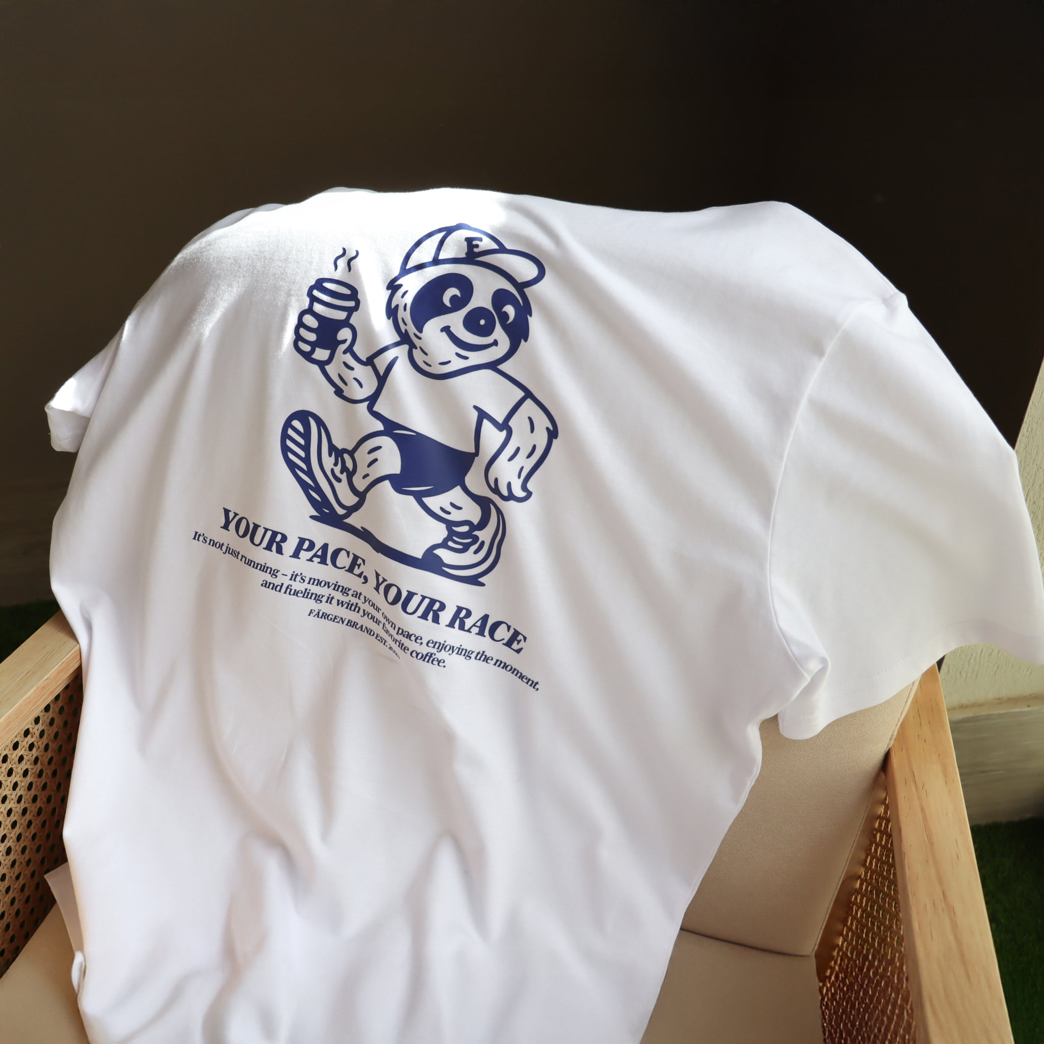 Camiseta Färgen Your Pace Your Race doblada sobre una silla mostrando el diseño trasero con perezoso corriendo y texto en azul.