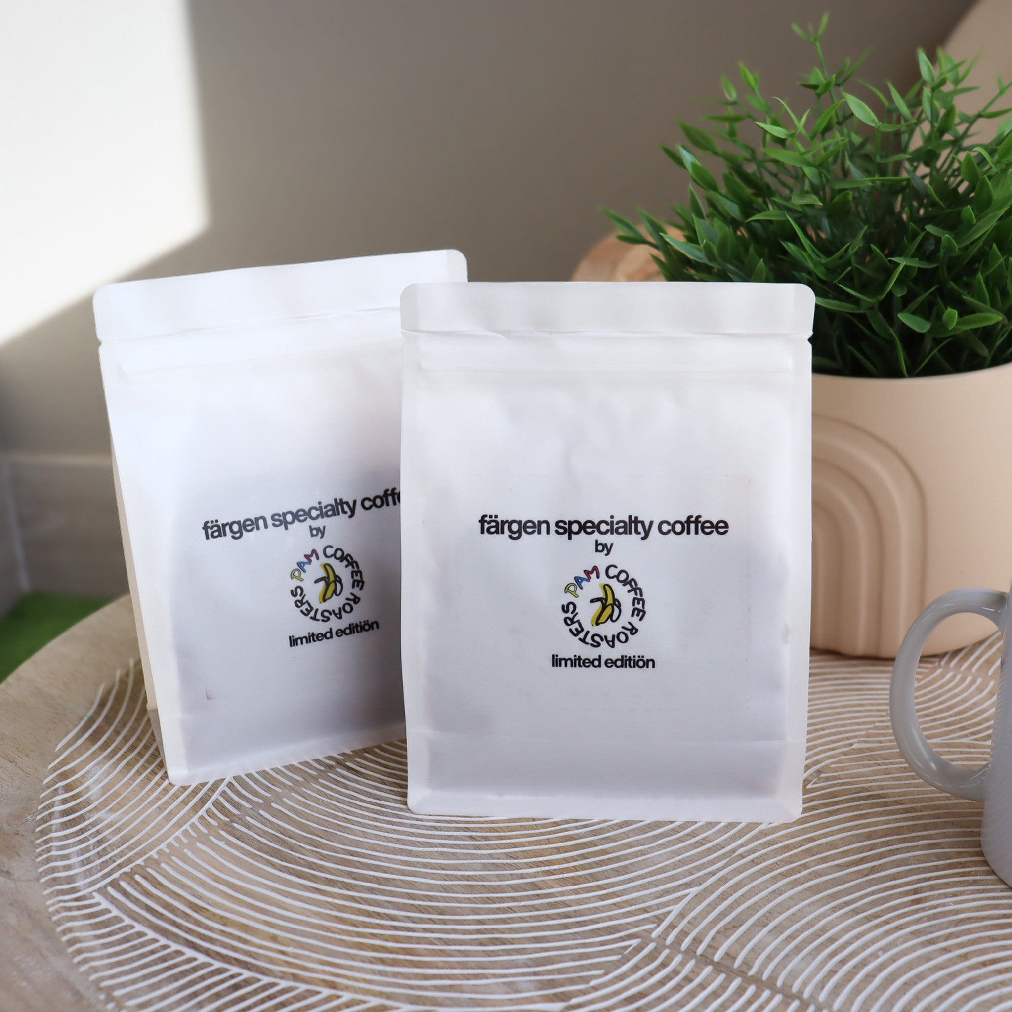 Dos bolsas de Färgen Specialty Coffee x Pam Coffee Roasters edición limitada junto a una planta y una taza, sobre mesa decorativa.