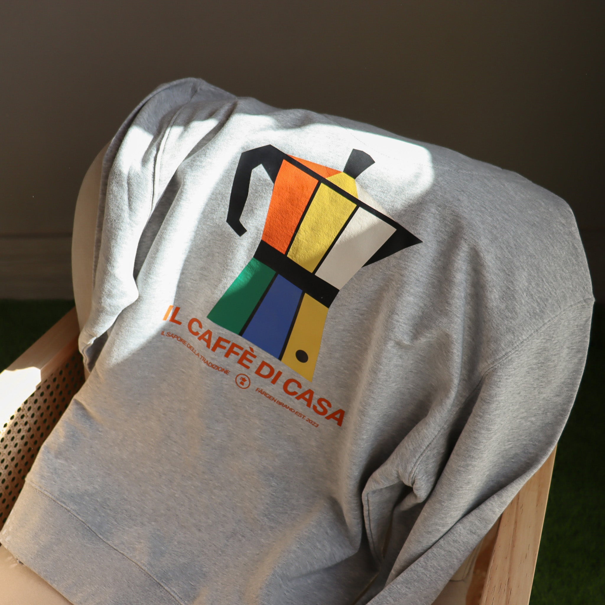 Sudadera gris Färgen Il Caffè di Casa doblada sobre silla mostrando el diseño trasero con cafetera italiana y texto Il Caffè di Casa.