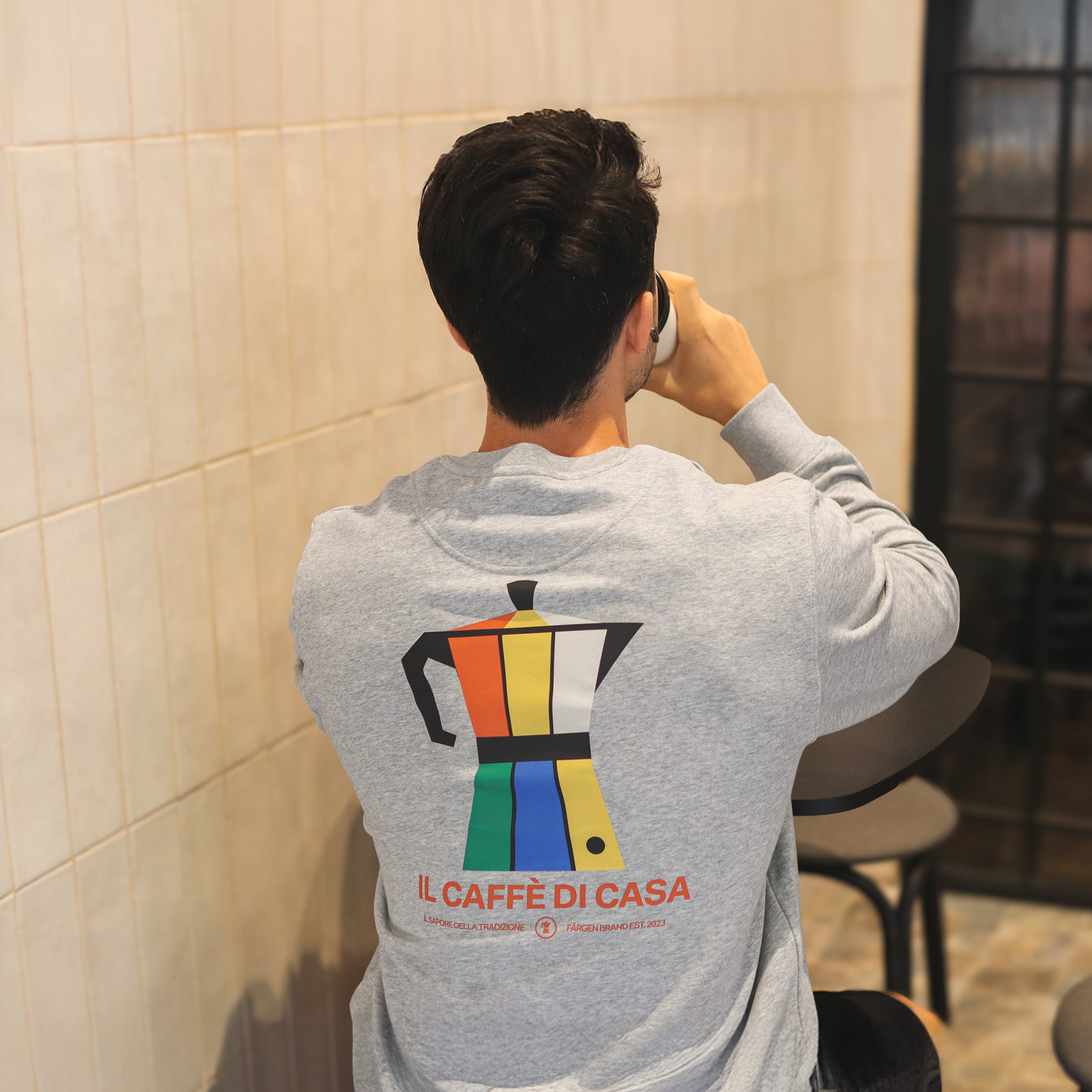 Modelo con sudadera gris Färgen Il Caffè di Casa de espaldas, mostrando el diseño trasero con cafetera italiana de colores Bauhaus y texto Il Caffè di Casa.
