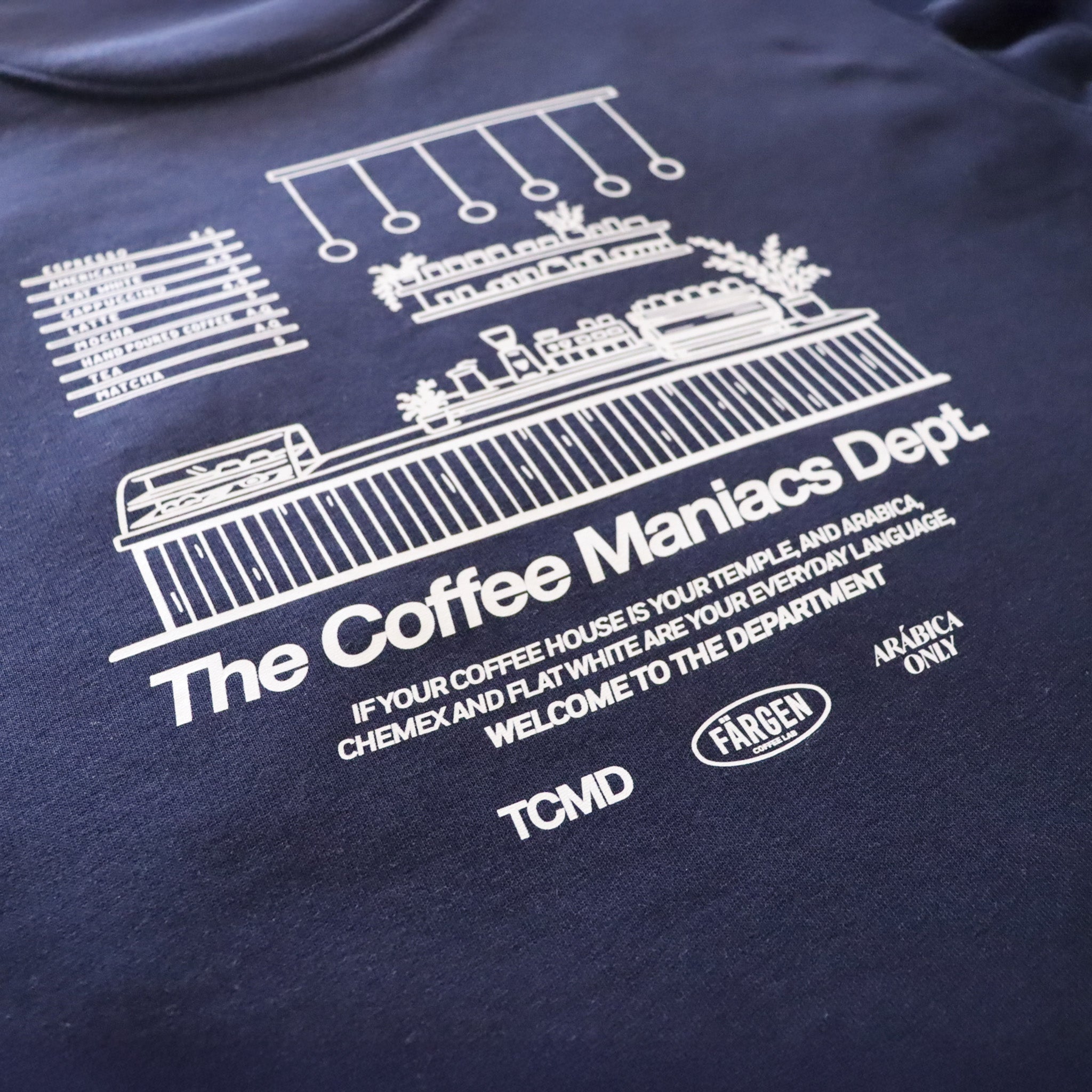 Detalle del diseño trasero de la sudadera Färgen The Coffee Maniacs Dept con ilustración de barra de cafetería y texto en blanco.