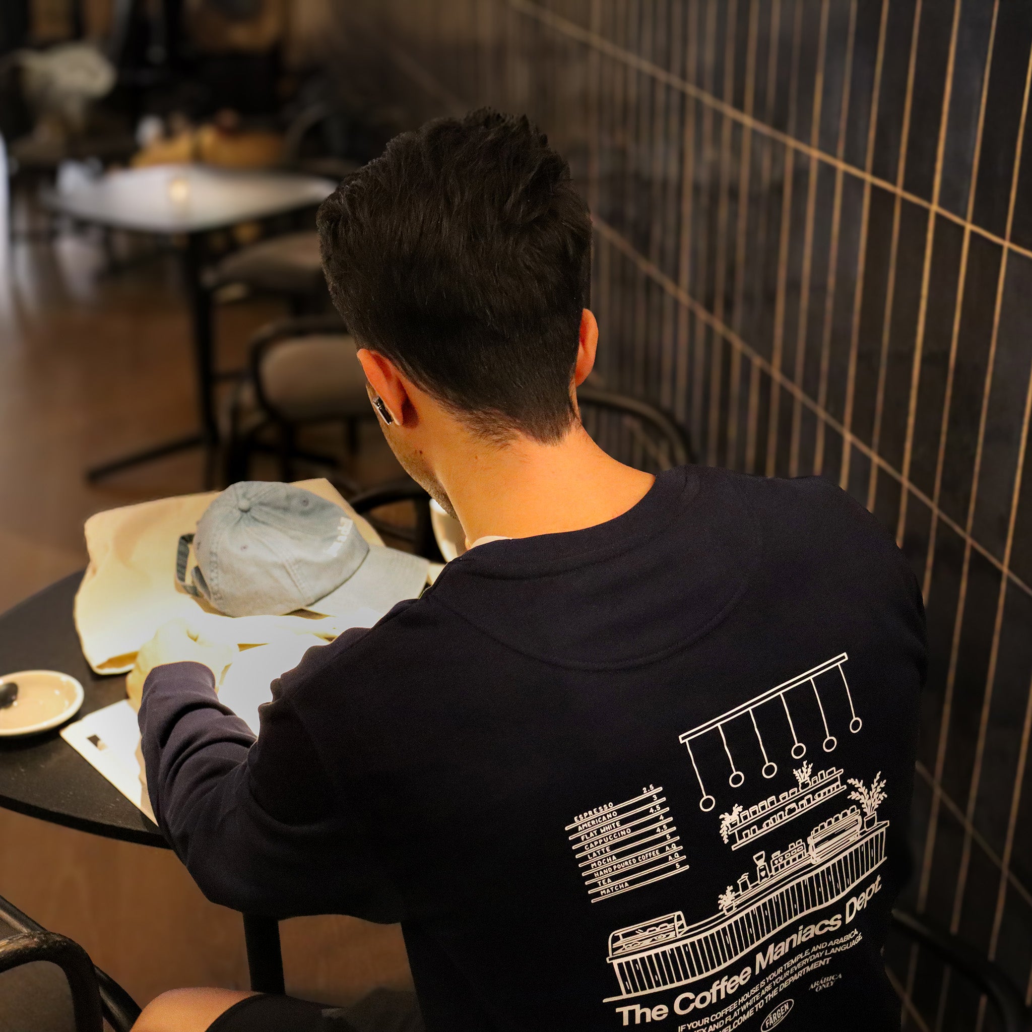 Hombre con sudadera azul marino Färgen The Coffee Maniacs Dept sentado en una cafetería mostrando el diseño trasero con ilustración de barra de café.