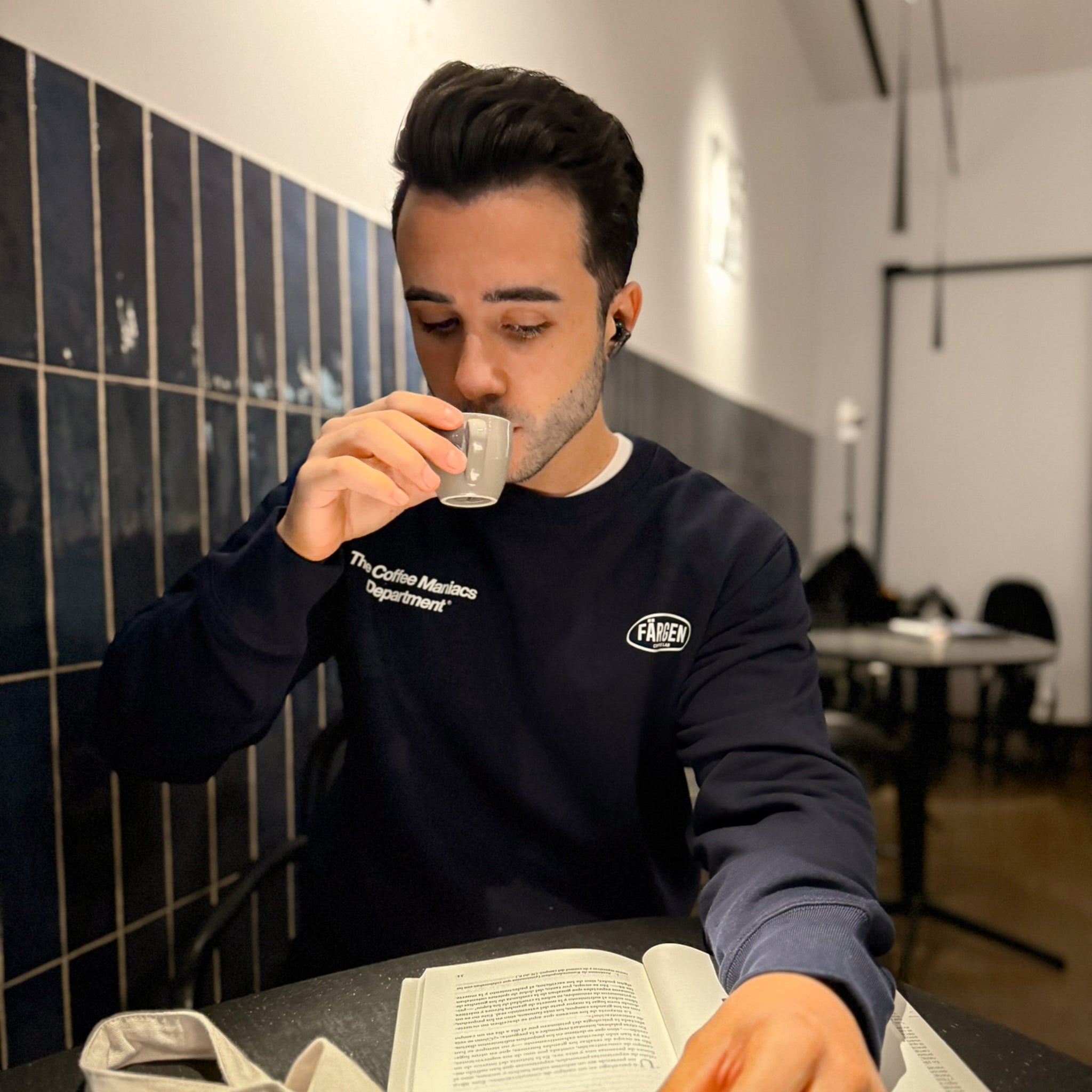 Hombre con sudadera azul marino Färgen The Coffee Maniacs Dept tomando café en una cafetería, mostrando el diseño delantero con logotipo Färgen Coffee Lab.